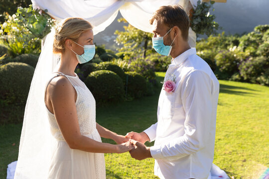 Happy caucasian bride and groom getting married wearing face masks and holding hands