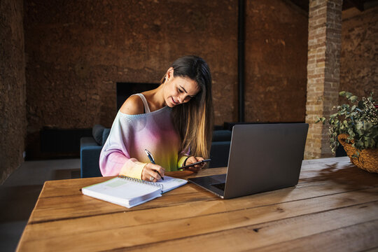 Smiling freelancer with smartphone and laptop writing in copybook indoors