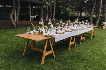 Wooden, long decorated with flowers, reeds, empty dishes, plates, forks, knives, glasses with a white tablecloth stands on the green grass in the park, forest. Wedding banquet.