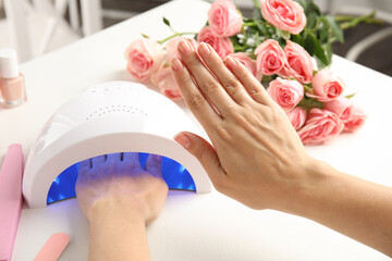 Woman using ultraviolet lamp to dry gel nail polish at white table, closeup