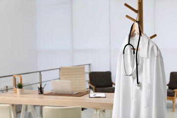 Modern doctor's workplace with wooden table and white coat hanging on rack in office - obrazy, fototapety, plakaty
