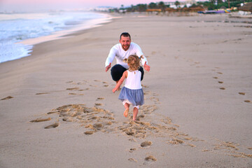 A little girl runs to her dad on the sand by the sea. The child is happy to travel with his parents. man meets girl beach running towards her father. open arms father on beach at the sunny day