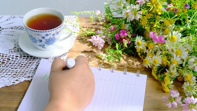 female hands write hello, a cup of tea, a beautiful summer bouquet of wildflowers, a handmade lacy white doily on the table, the concept of summer time, a tea party in the garden
