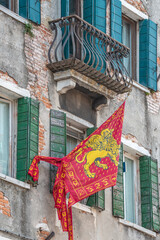 Old traditional city flag of Venice, Italy, depicting Venetian lion with wings and Bible hanging at very old deteriorated building facade.