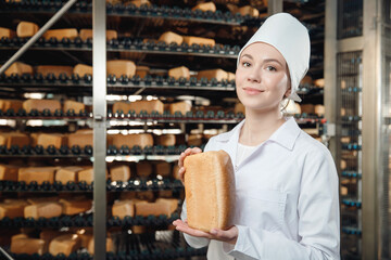 Woman baker holding fresh bread in hands on background automatic conveyor of bakery food factory - obrazy, fototapety, plakaty
