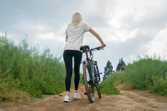 Young slender blonde woman in a white shirt, black leggings and white sneakers rolls a bicycle along a sandy road in the middle of a field on a cloudy summer day, photo from the back