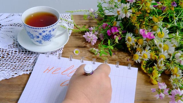 female hands write hello august, a cup of tea, a beautiful summer bouquet of wildflowers, a handmade lacy white doily on the table, the concept of summer time, a tea party in the garden