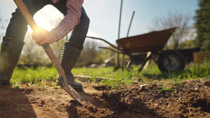Male farmer in plaid shirt and straw hat is digging in garden with shovel at sunny spring day shot...