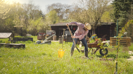 Male farmer in plaid shirt and straw hat is digging in garden with shovel at sunny spring day shot in 4k super slow motion