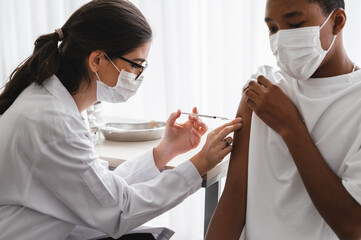 A female doctor or nurse wearing a mask and face shield is injecting the coronavirus 19 vaccine on the shoulder of an African-American man to immunize. Concept of preventing the spread of COVID-19.