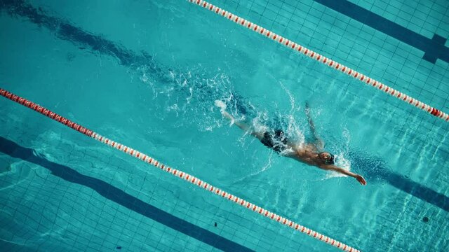 Aerial Top View: Muscular Male Swimmer in Swimming Pool. Professional Athlete Swims in Backstroke Style, Determination in Training to Win Championship. Cinematic Spinning Shot with Stylish Colors