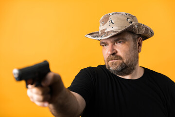 Middle aged man with gun in black t-shirt and hat against yellow background.