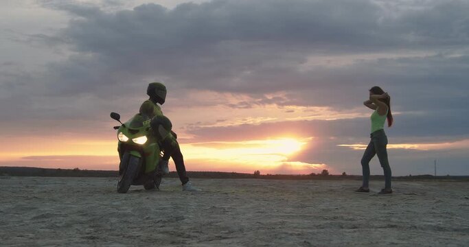 In a vacant lot, a motorcyclist leaned on his sports bike. A beautiful girl in a green T-shirt approaches him, hugs and kisses him on the helmet. In the background, the sun sets behind the clouds