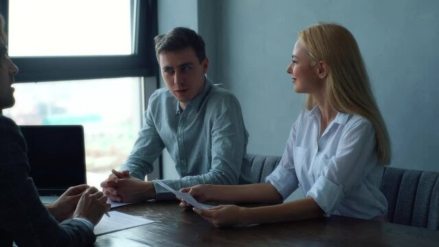 Caucasian HR male and female managers reading resume and interviewing businessman sitting at desk near window. Smiling young woman and man headhunters holding job interview with young man candidate.