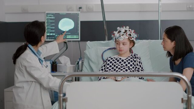 Pediatrician physician woman doctor discussing disease evolution expertise while monitoring brain tomography during recovery consultation. Sick child wearing eeg brain sensors headset in hospital ward