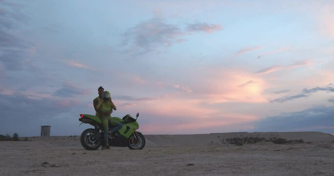 Athletic and handsome man and woman are standing near a high-speed motorcycle in nature. The guy hugs his girlfriend. She is wearing a protective helmet. Behind them is the colorful sky