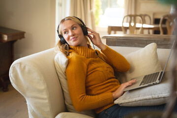 Obraz premium Smiling caucasian woman relaxing on couch in living room wearing headphones and using laptop
