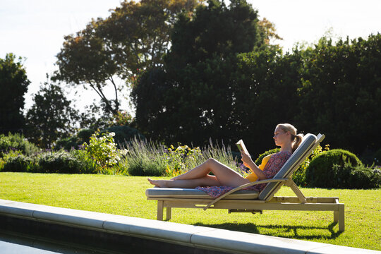 Smiling caucasian woman relaxing on sun lounger in beautiful sunny garden reading book