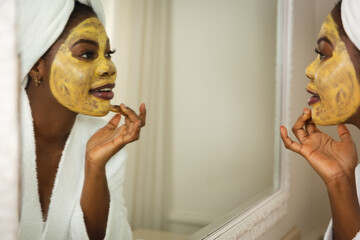 Happy african american woman in bathroom with towel on head, looking in mirror applying face mask