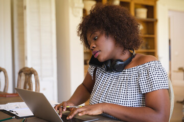 African american woman working in living room using laptop and talking on smartphone