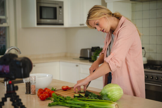 Caucasian woman in kitchen chopping vegetables and using camera, making cooking vlog - Powered by Adobe
