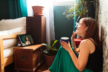 Young woman making a video call sitting at home and drinking coffee on the floor.