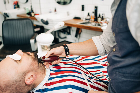 Crop barber cutting beard of client with trimmer in barbershop