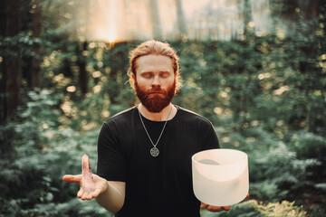 Red-haired man holding a singing bowl and a magic crystal in the forest Spiritual music meditation - 442753777