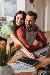 Fototapeta premium Woman embracing smiling boyfriend near newspaper, notebook and laptop