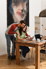 Smiling man pointing at laptop while girlfriend working near coffee at home
