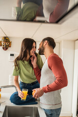 Cheerful man touching chin of girlfriend with glass of orange juice in kitchen