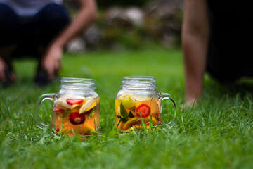 Two Jars of glass, delicious detox drink with red, orange and yellow fruits, standing on the grass. Selective Focus Glasses.