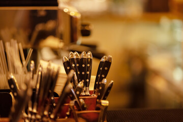 Cutlery stands in a wooden stand in a kitchen, cafe, restaurant, bistro in warm light. Spoons forks knives with black handle with a round pattern