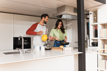 Obraz premium Smiling man holding jug with orange juice near girlfriend cooking pancakes