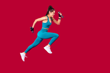 Athletic young girl in sports uniform and white snickers makes lunges with dumbbells in her hands over the red background.