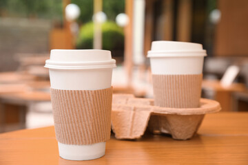 Takeaway paper coffee cups with plastic lids, sleeves and cardboard holder on wooden table outdoors