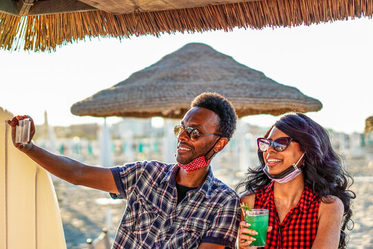 African american couple taking selfie in beach party - Young friends with face mask drinking cocktail in holiday