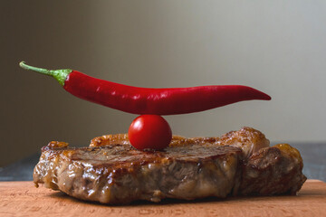 Grilled meat piece of ribeye beef meat on wooden cutting board on a wooden table stacked tomatoes garnished levitating hot pepper. Fresh food. BBQ.