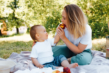 Cute toddler son feeding fruit to his young mother, outdoors. Family picnic in nature, mom and baby are resting on a blanket on the green grass outside. Beautiful mother and child eating peach.