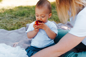 Little kid is funny eating a ripe peach, sitting on a blanket on the green grass, outdoors. Family picnic in the summertime. Healthy and wholesome food for the child.