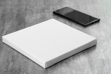 White empty square printable canvas and black phone next to it on gray table. Mockup canvas (944)