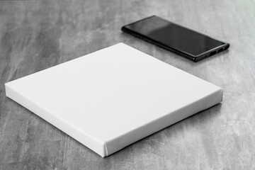 Fototapeta premium White empty square printable canvas and black phone next to it on gray table. Mockup canvas (277)