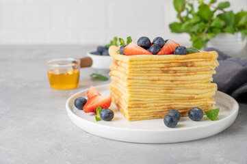 Stack of homemade crepes served with fresh blueberries and strawberries on a white plate on a gray concrete background. Food for Maslenitsa. Copy space.