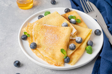 Homemade crepes served with fresh blueberries and powdered sugar on a white plate on a gray concrete background. Food for Maslenitsa. Copy space.