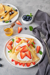 Crepes or thin pancakes with cream cheese, fresh strawberries and honey on a white plate on a gray concrete background. Copy space.