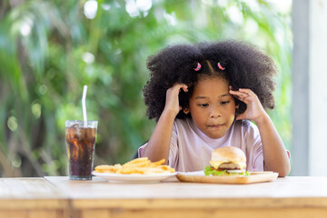 A little curly haired African American girl sits at the table looking at hamburgers and french fries, thinking about whether to eat. unhealthy food Childhood concepts and eating.