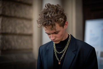 Positive male teenager wit curly hair dressed in street style clothes with chains around neck for a...