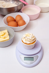 Freshly prepared cupcake with butter cream on the scales, in the background are the ingredients for its preparation. Selective focus.