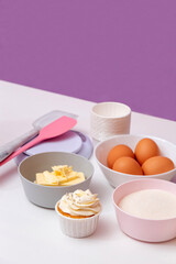 Sugar, eggs, butter, spatulas, scales, molds for making cupcakes with freshly prepared cake on a white table. Purple background with space for text. Selective focus.
