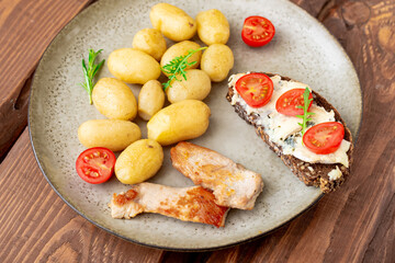 Boiled young potatoes, chunks of grilled turkey meat and cereal bread with blue cheese and tomatoes on a plate on a wooden background.
